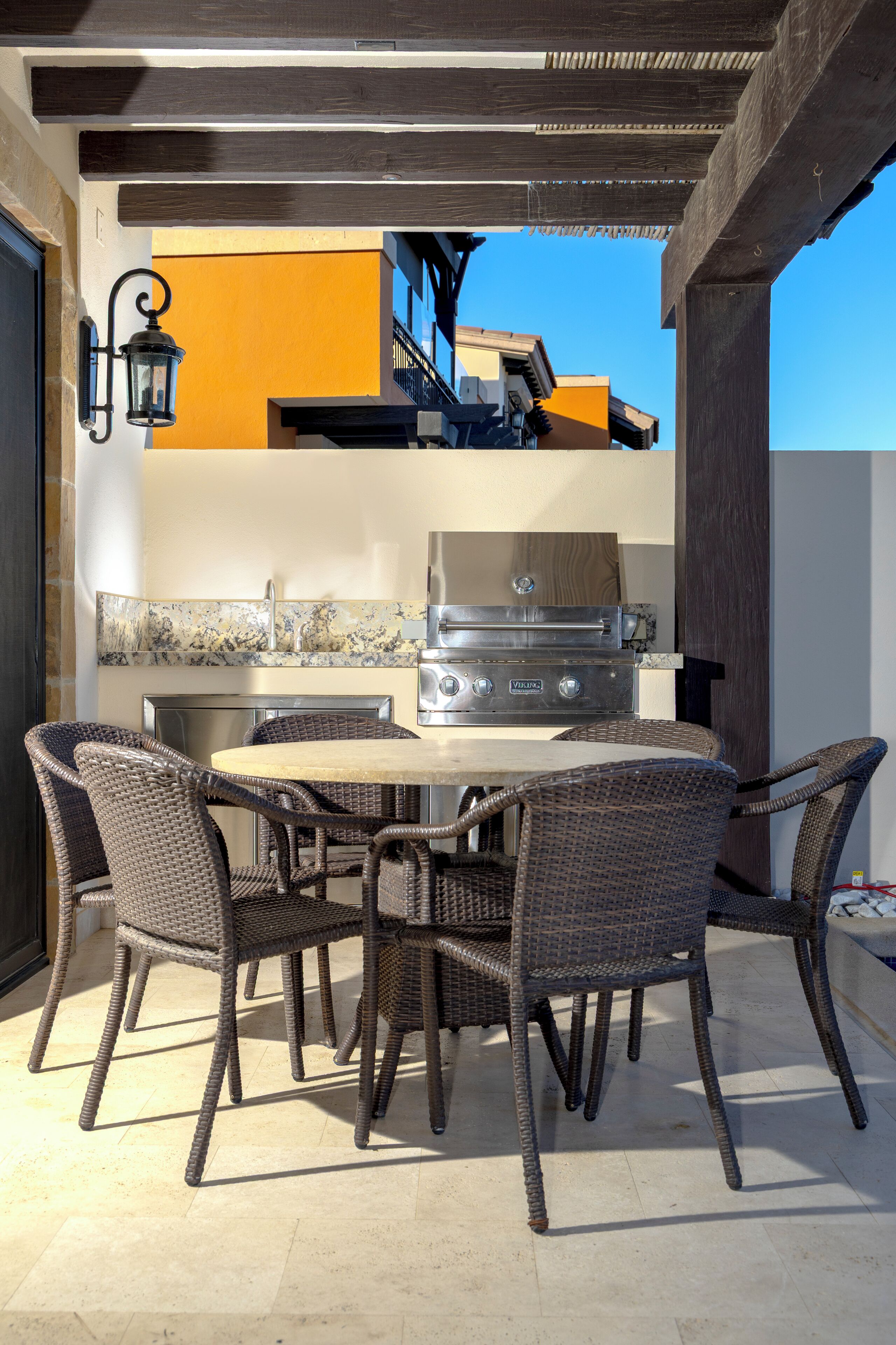 Un patio exterior con una mesa redonda y sillas de mimbre, una parrilla y un farol fijado a la pared.