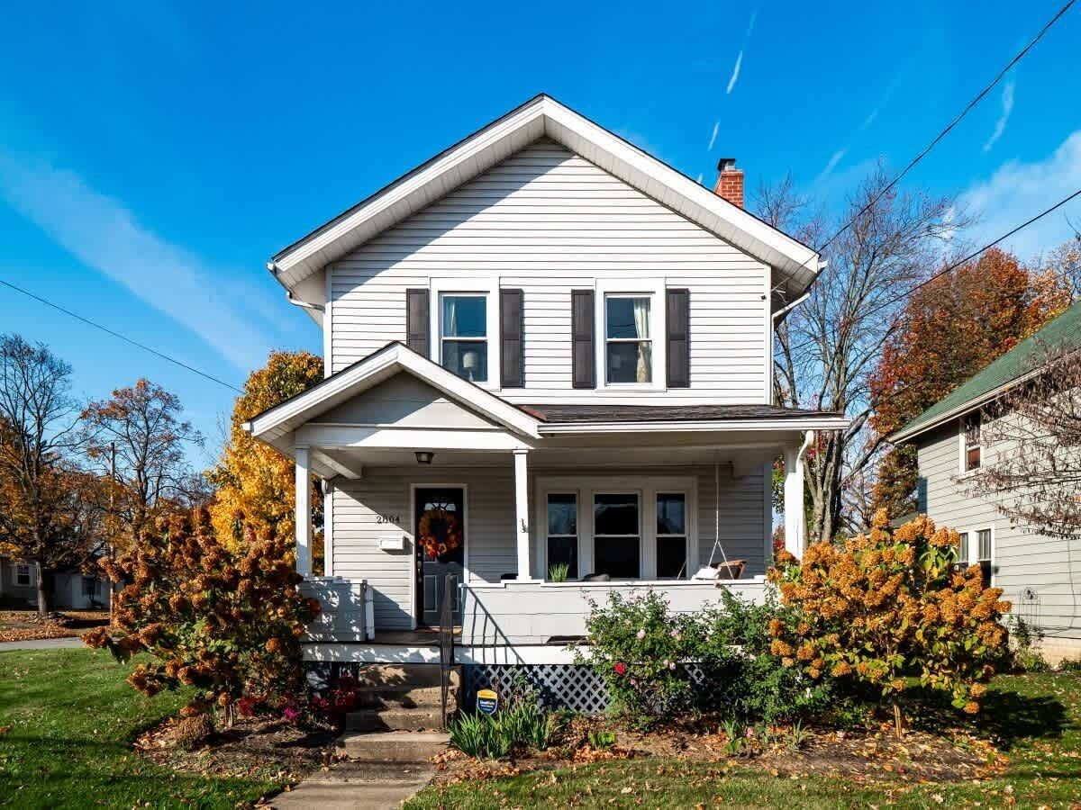 A two-story house with a front porch, a visible front door, and a small garden area with shrubs and a lawn.