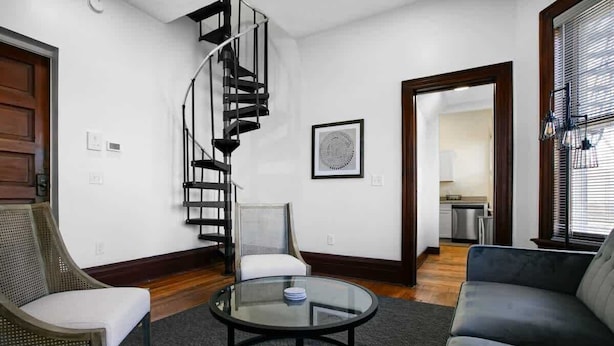 Una sala de estar moderna con una escalera espiral, una mesa de centro redonda de vidrio, un sofá gris y una silla de mimbre.