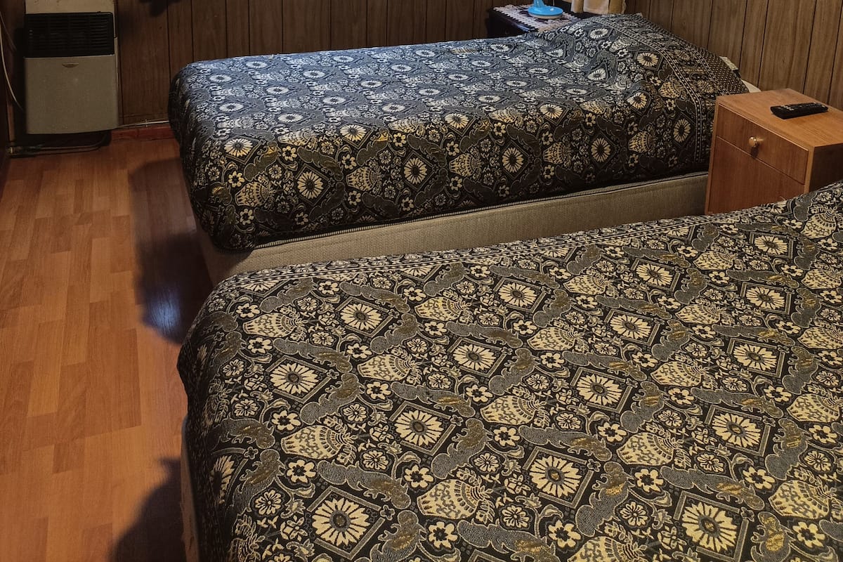 A room with two beds, wooden walls, a window with curtains, a small table, and an air conditioning unit.
