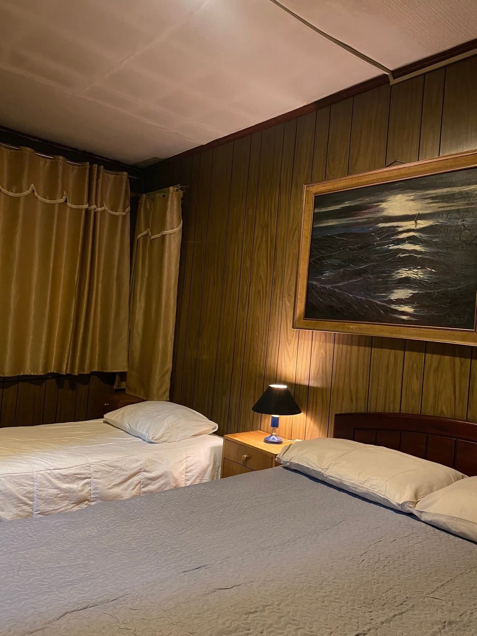 A hotel room with two beds, a nightstand with a lamp, wooden paneling, and a painting of a landscape.