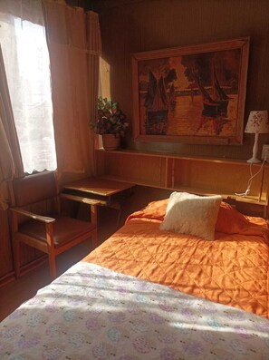 Una habitación con cama, escritorio, silla, ventana con cortinas, un cuadro en la pared y una lámpara.
