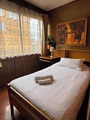 Une chambre d’hôtel avec un lit, un tableau au mur, une fenêtre avec des rideaux et une table de chevet avec une lampe.