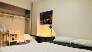 Una habitación de hotel con una cama, un escritorio, una silla, un ventilador de pie y una lámpara de pared.