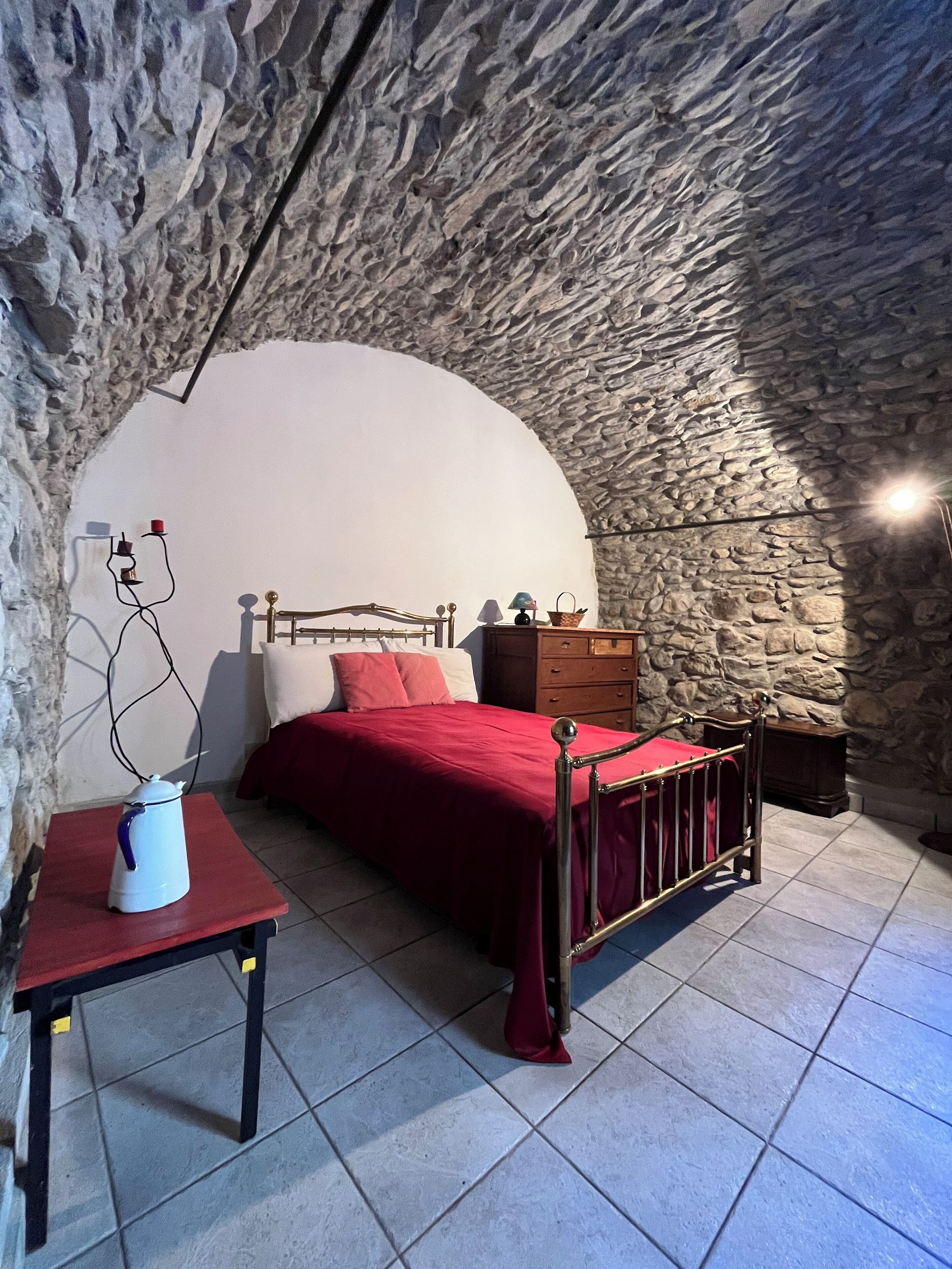 Une chambre avec un mur en pierre, un lit recouvert d’une courtepointe rouge, une commode en bois et une petite table avec une bouilloire.