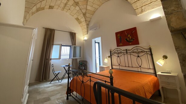 A bedroom with a stone ceiling, a bed with an orange bedspread, a nightstand, a chair, and a painting on the wall. - HOUSE CENTER CASTRIGNANO 3 (CASTRIGNANO DEL CAPO)