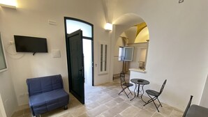 A room with a blue sofa, a small table with two chairs, a TV mounted on the wall, and a door leading to another room. - HOUSE CENTER CASTRIGNANO 2 (CASTRIGNANO DEL CAPO)