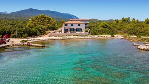 Una propiedad costera con un mar turquesa cristalino, un edificio de dos pisos con tejado de tejas y montañas al fondo.
