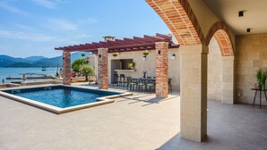 Zona de piscina con pérgola, mobiliario y vista al mar.