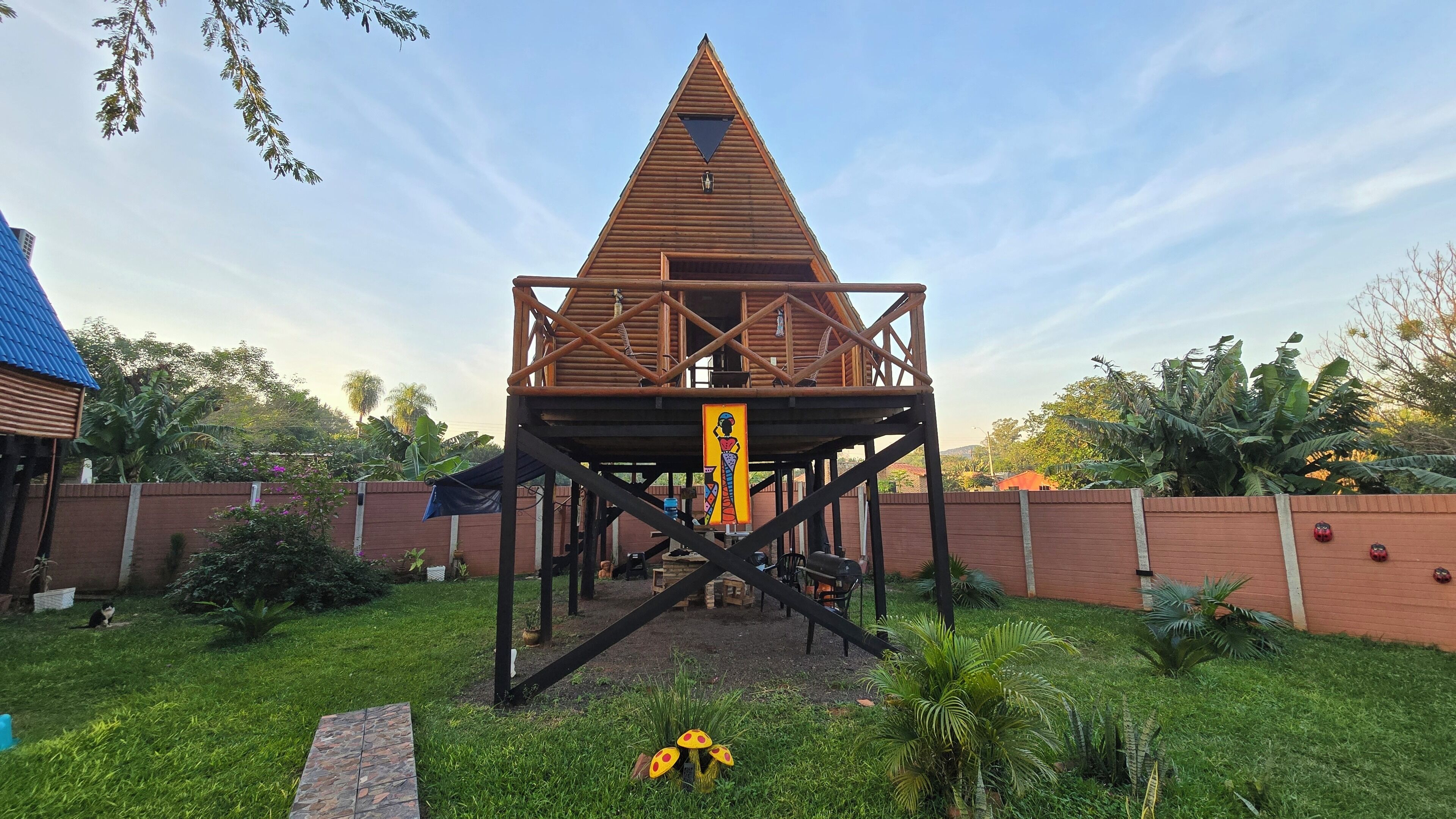 Une cabane en bois avec un toit triangulaire et une échelle menant à l’intérieur, entourée d’un mur de briques et de verdure.