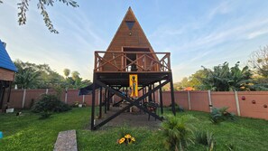 A wooden playhouse with a triangular roof and a ladder leading up to it, surrounded by a brick wall and greenery. - Alpinas Kamba (Paraguari)