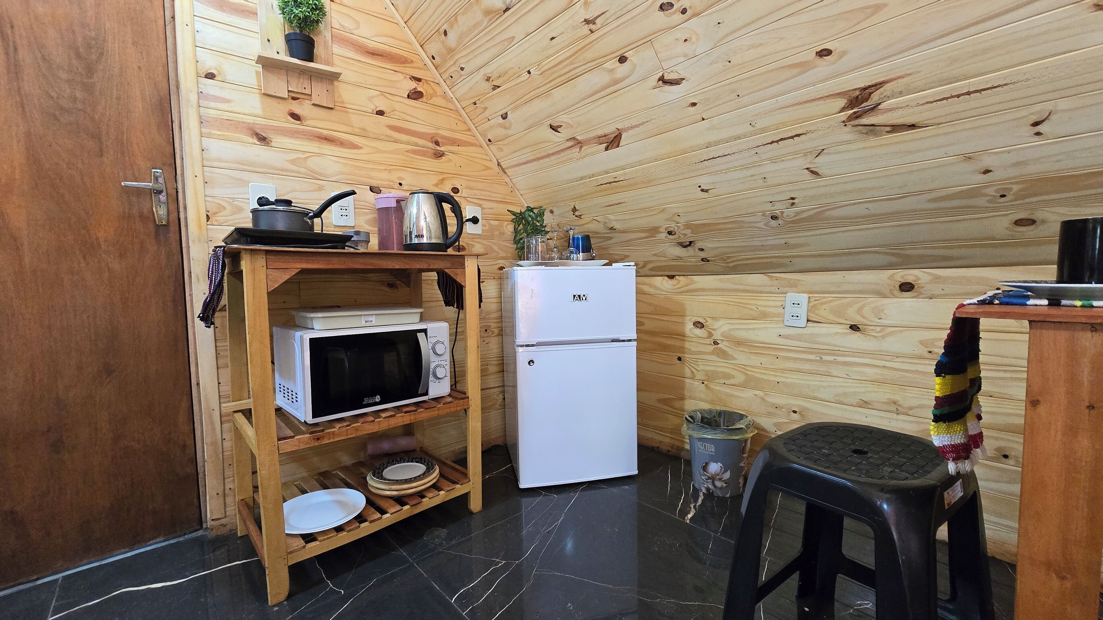 A compact kitchen with wooden walls, a white refrigerator, a microwave, and a small shelf with kitchenware.