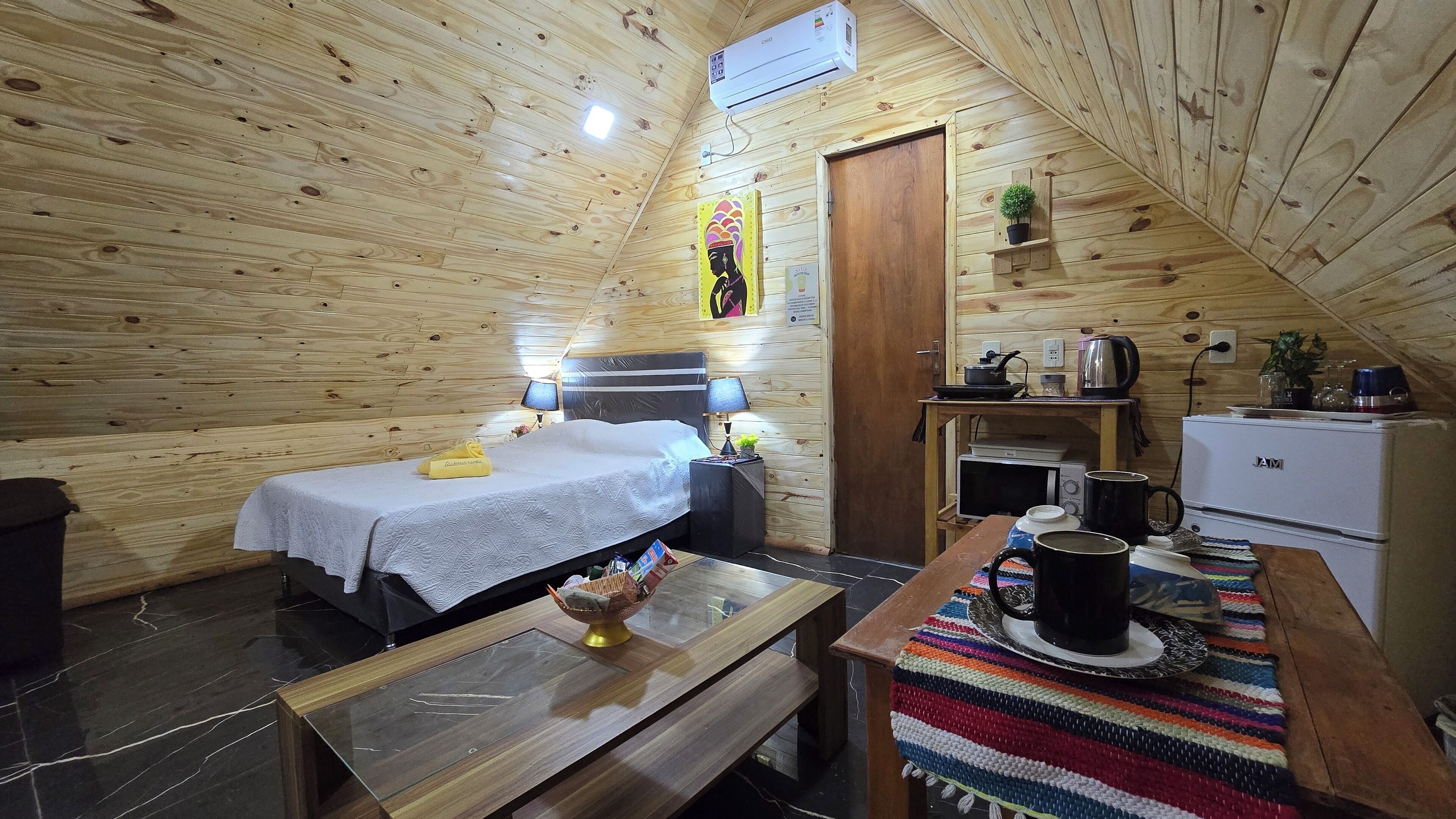 A cozy wooden cabin room with a bed, bedside table, lamp, small table with a basket, and a kitchenette area with a fridge, microwave, kettle, and coffee maker.
