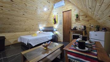 A cozy wooden cabin room with a bed, bedside table, lamp, small table with a basket, and a kitchenette area with a fridge, microwave, kettle, and coffee maker.