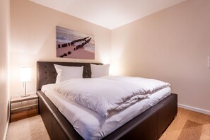 Ein ordentlich bezogenes Bett in einem Hotelzimmer mit Kopfteil, einem Nachttisch mit Lampe und einem Wandbild, das eine Küstenlandschaft zeigt.