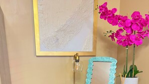 A green cabinet with two gold handles, a yellow chair, a framed mirror, and a potted orchid. - Vibrant Furnished Studio, Walk to LSUNO, 30+ Day Stays (New Orleans)