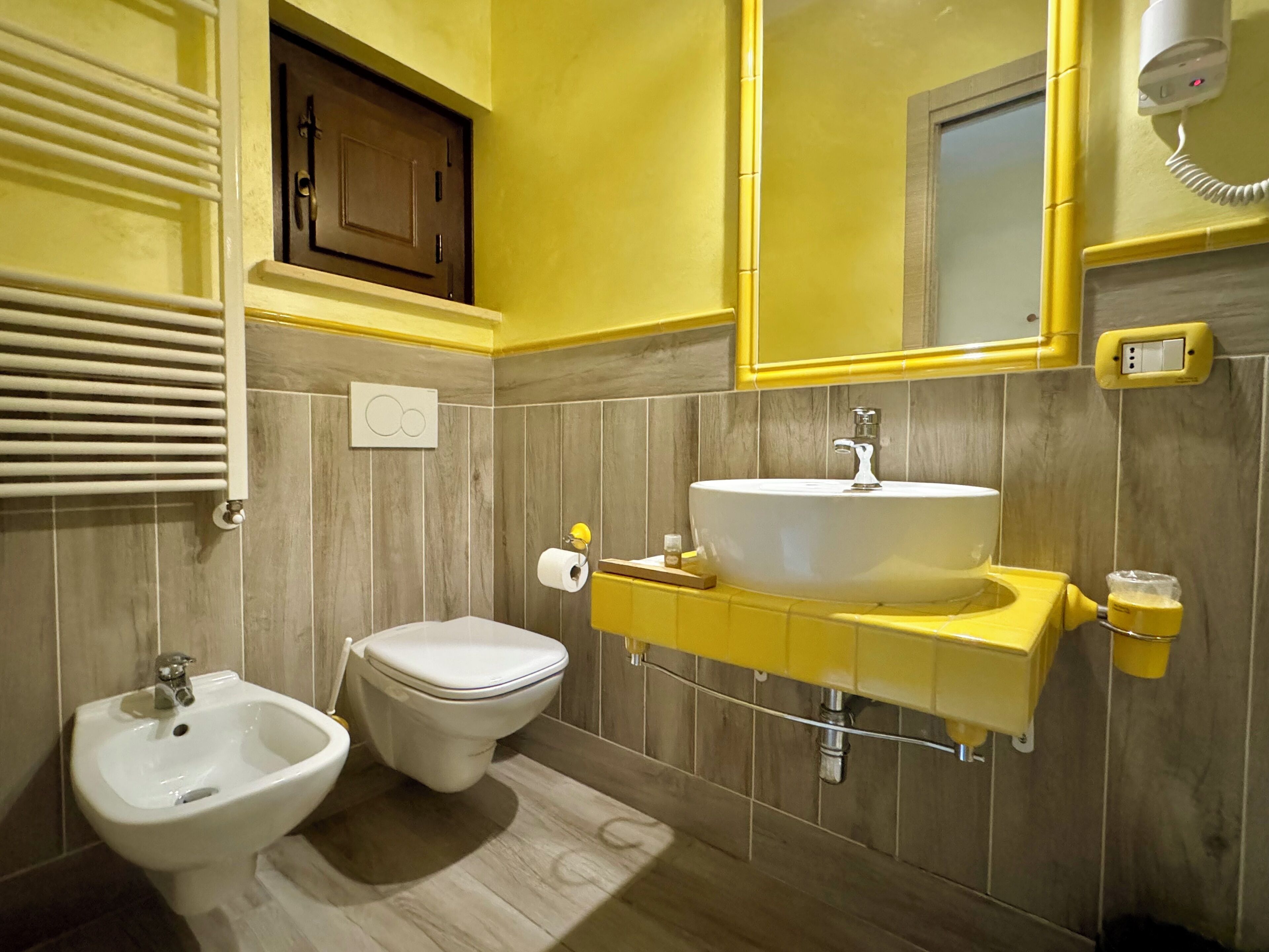 Une salle de bain avec un lavabo blanc, des toilettes et un bidet. Un miroir est placé au-dessus du lavabo et un porte-serviettes est fixé au mur.