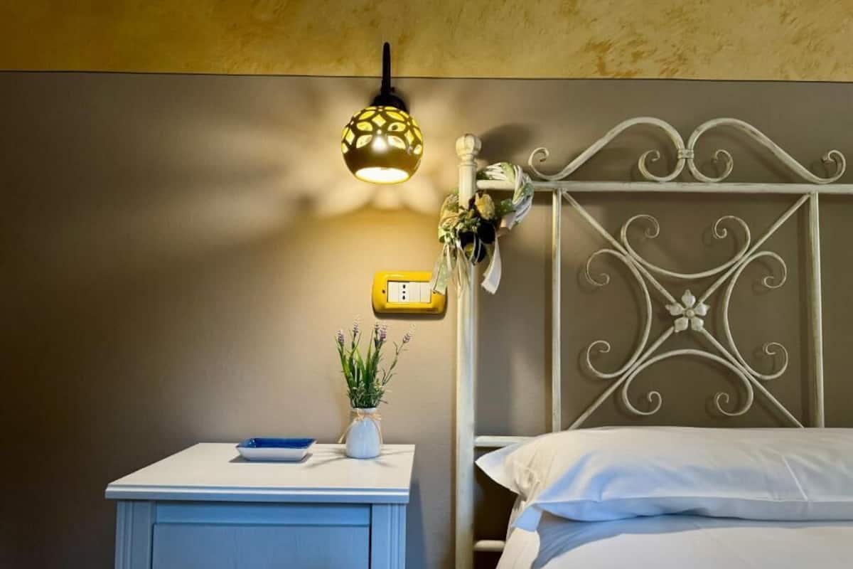Ein Schlafzimmer mit einem weißen Metall-Kopfteil, einem Nachttisch mit einer blauen Ablage und einer weißen Vase mit Blumen, sowie einer wandmontierten Leuchte.