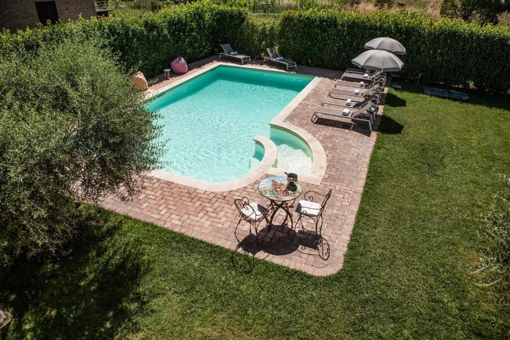 A rectangular swimming pool with a brick border, surrounded by a grassy lawn and hedges. There are lounge chairs and a table with chairs near the pool.