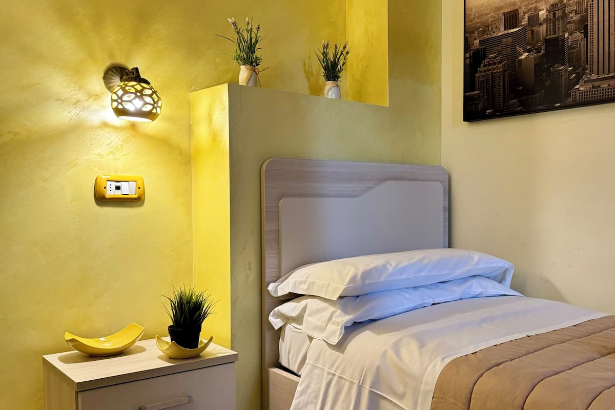 Ein Schlafzimmer mit Bett, Nachttisch und einem Beistelltisch mit Lampe. Auf dem Beistelltisch stehen zwei Blumentöpfe.