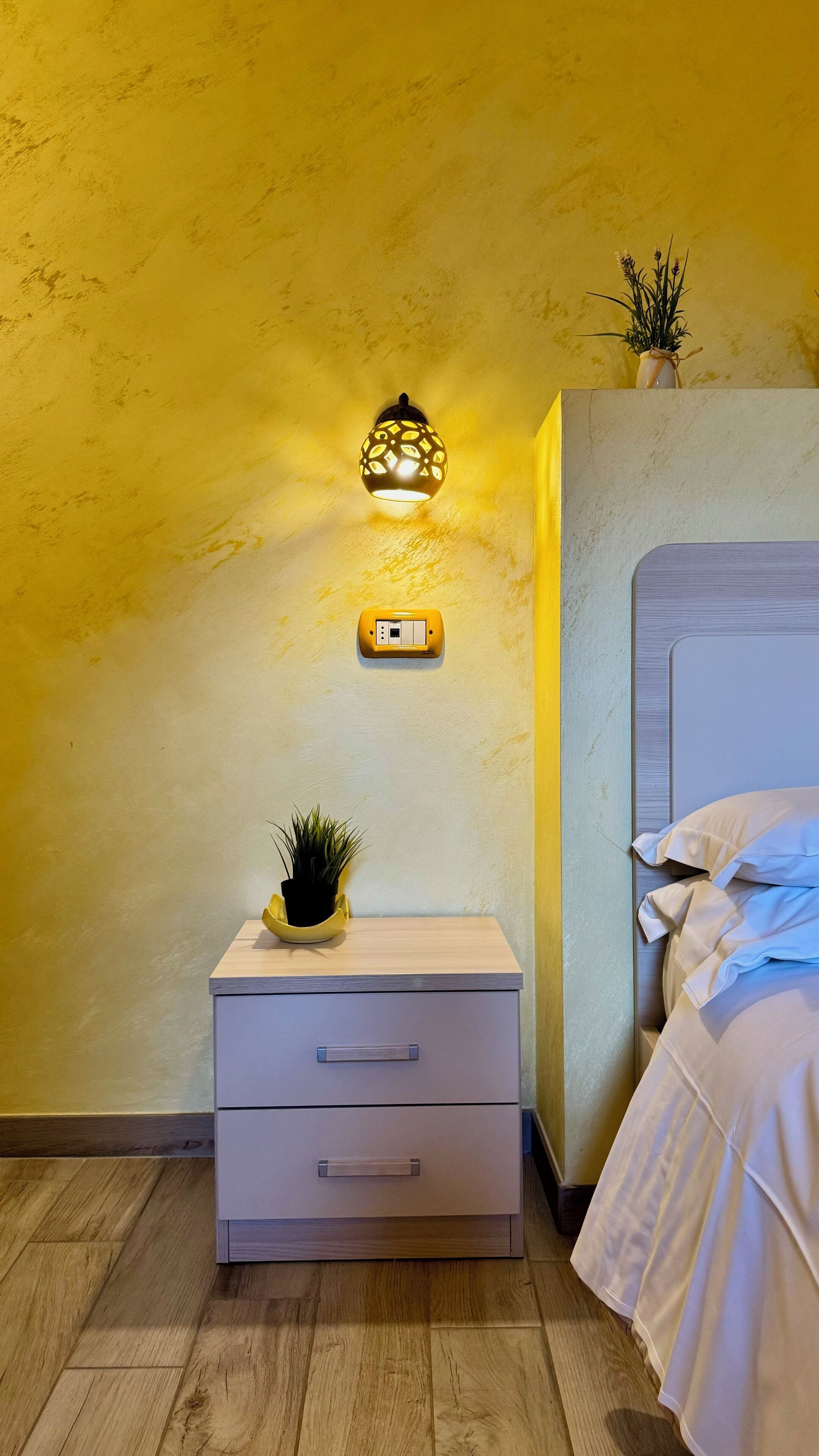 Een slaapkamer met een bed, een nachtkastje met lamp en een wandcontactdoos.