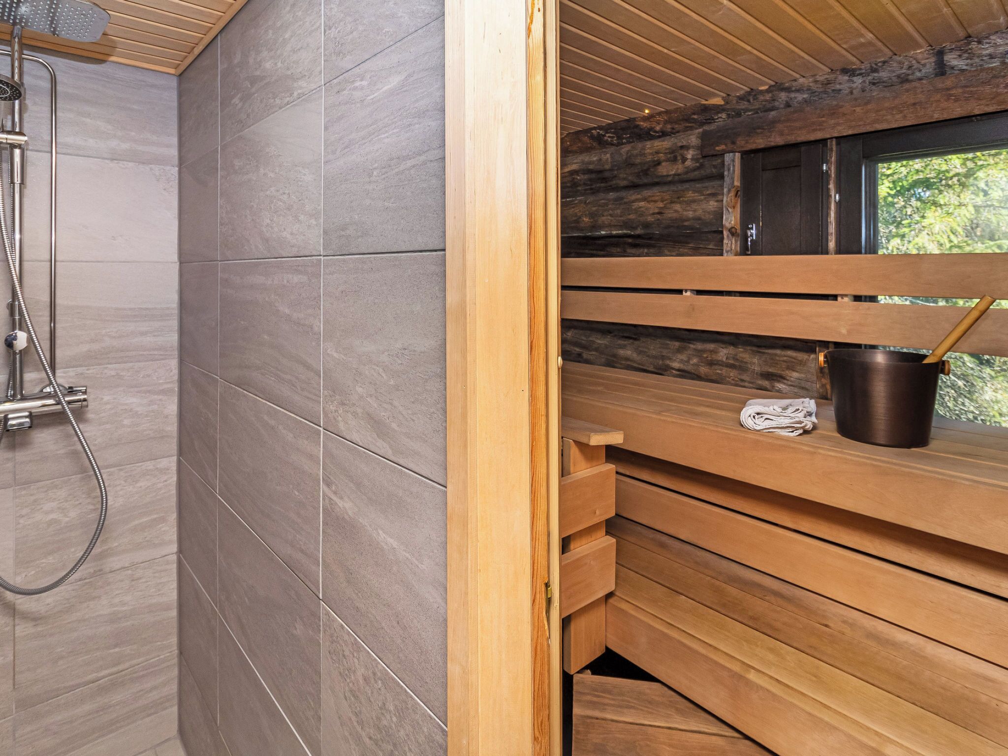 A wooden sauna with a bench, a shower area with grey tiles, and a window overlooking greenery.