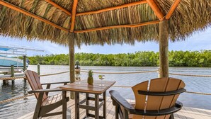 A wooden deck with a thatched roof, a table with chairs, and a view of a river and greenery.