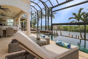 A luxurious indoor pool area with wicker furniture, a glass ceiling, and a view of a natural water body. - WOW!! Experience Luxury! Gulf Access, Pool & Spa, Kayaks, Dock, Tiki- Villa Waterfront Wonderland (Cape Coral)