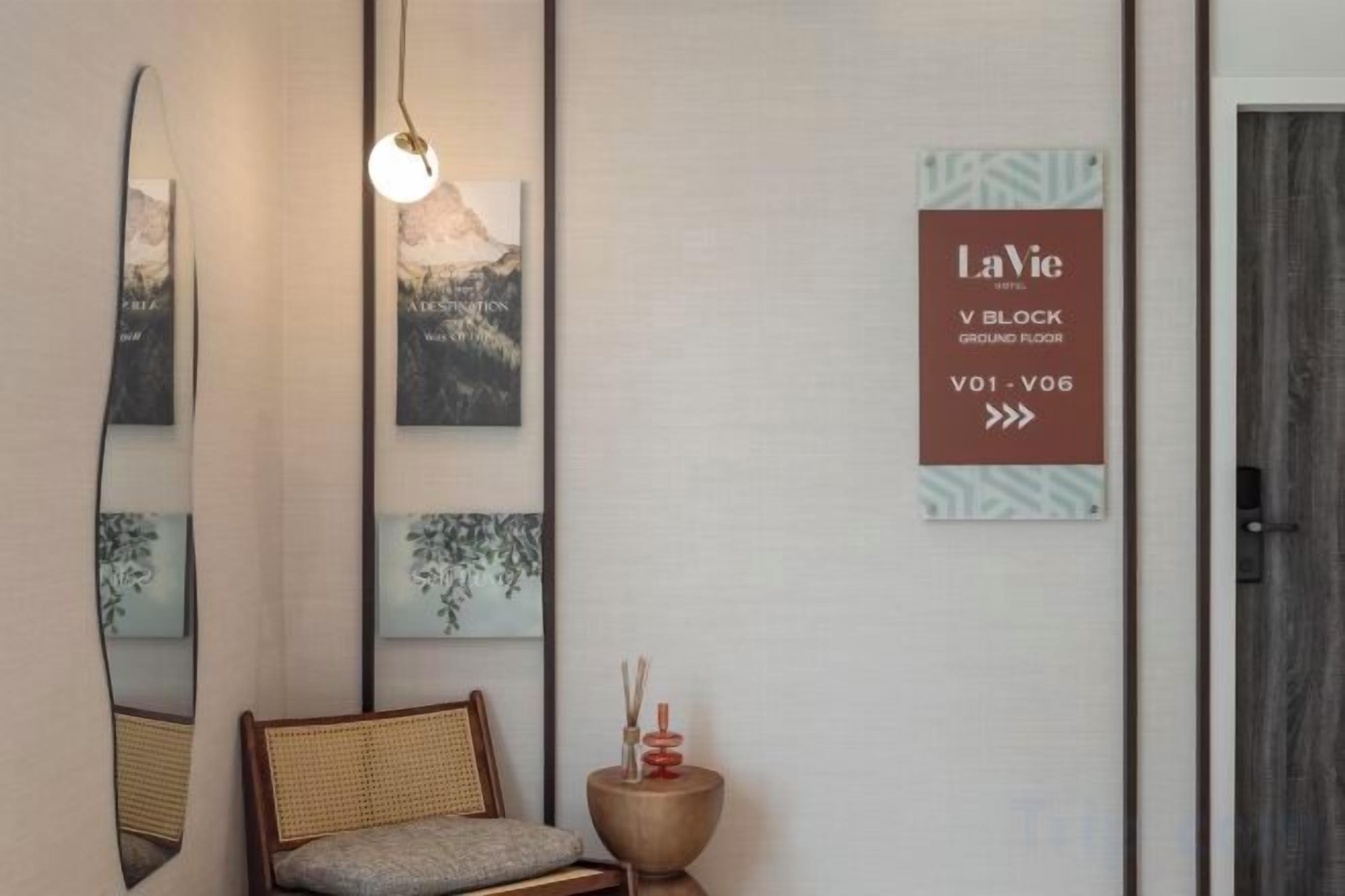 A reception area with a chair, a round side table with candles, and wall-mounted artwork. A sign indicates the location as 'L'Avic V Block Ground Floor.'