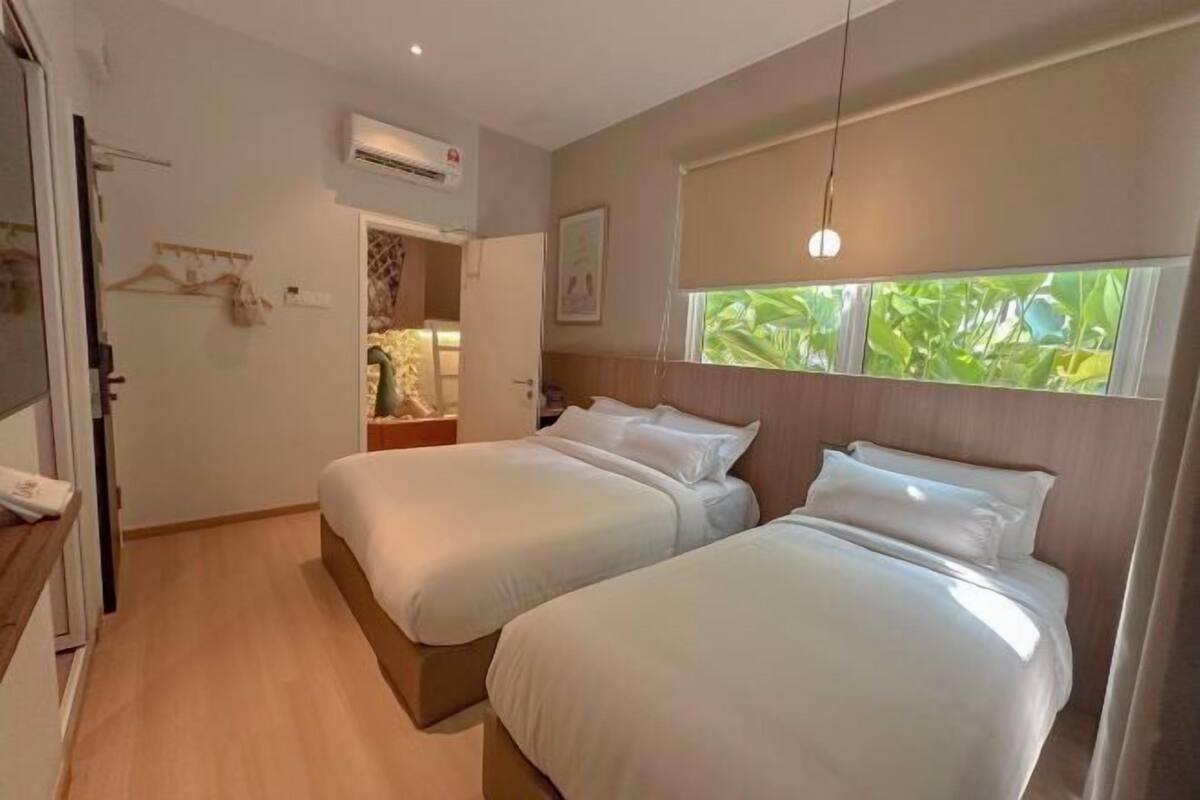Une chambre d’hôtel avec deux lits, un plancher en bois, un appareil de climatisation, une fenêtre donnant sur un espace verdoyant et un luminaire suspendu.