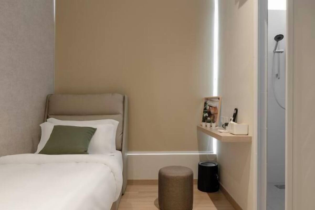 Une chambre d’hôtel moderne équipée d’un lit, d’un petit tabouret, d’une étagère avec des articles de toilette et d’un espace douche.