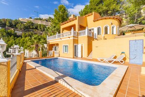 Una piscina con un solarium in legno, un balcone bianco e vista su una zona montuosa.