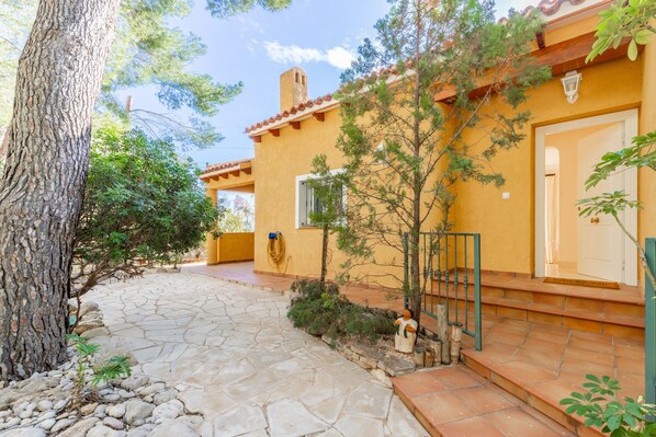 Un sendero de piedra conduce a un edificio amarillo con una puerta y una ventana, rodeado de árboles y una pequeña estatua.