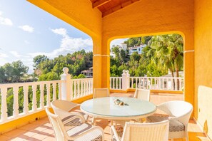 Zona relax esterna coperta, con un tavolo rotondo e sedie in vimini, affacciata su un paesaggio verdeggiante con alberi e edifici.