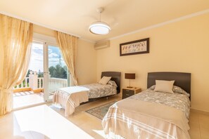 Una camera d'albergo con due letti, un balcone, una ventola a soffitto e un quadro appeso al muro.