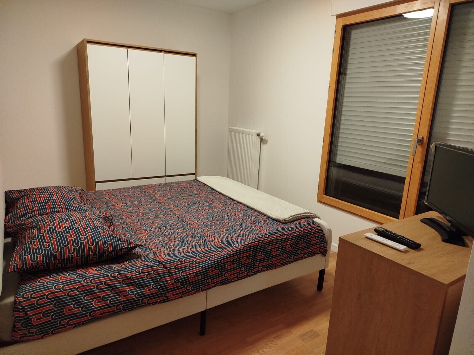 A bedroom with a bed, a desk with a computer, a window with blinds, and a wardrobe.