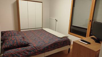 A bedroom with a bed, a desk with a computer, a window with blinds, and a wardrobe.