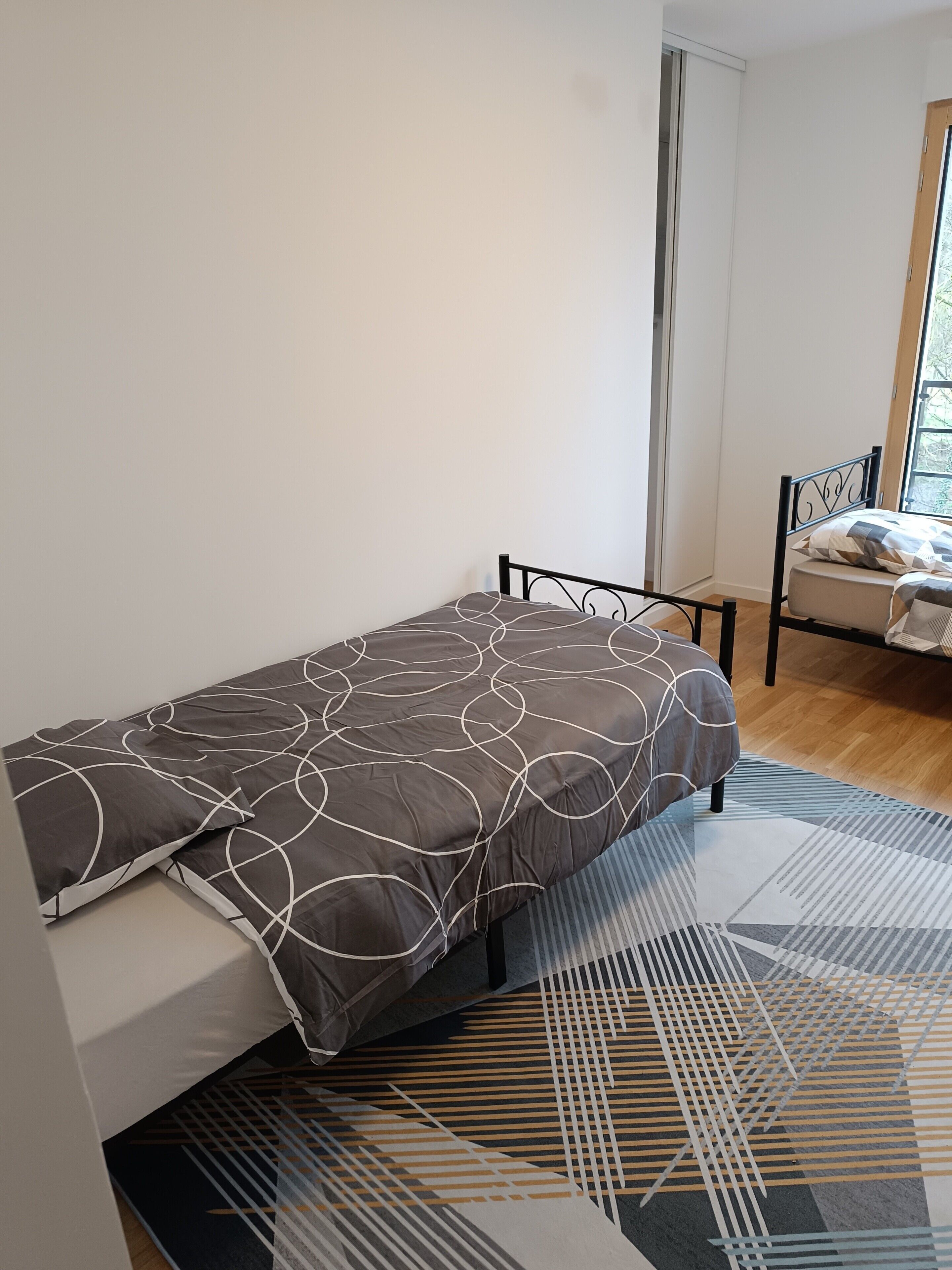 A single bed with a patterned bedspread, a metal headboard, and a geometric rug on a wooden floor.