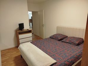 A bedroom with a bed, a bedside table with a TV, a wardrobe, and a door leading to another room. - Apartment 'Chez Sibylle Chevreuse Saclay' with Balcony, Wi-Fi and Air Conditioning (Saint-Rémy-lès-Chevreuse)