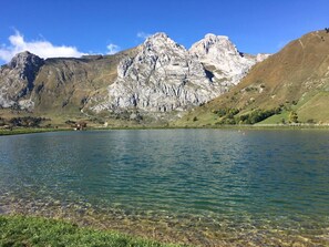Paisaje montañoso con un lago cristalino, terreno rocoso y un edificio pequeño cerca de la orilla.