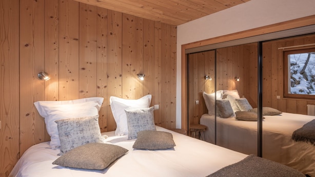 Une chambre avec des murs en bois, un grand lit avec de nombreux oreillers, une petite table et une fenêtre donnant sur des arbres.
