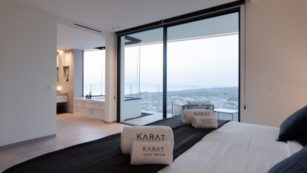 Ein modernes Hotelzimmer mit einem großen Bett, zwei Handtüchern mit dem Schriftzug 'Karat Luxury Rentals', einem Balkon mit Stadtblick und einem separaten Badebereich.