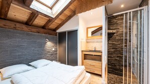 A bedroom with a skylight, a bed with white bedding, a wooden headboard, a dark wall, a bathroom with a stone wall, and a glass shower enclosure.