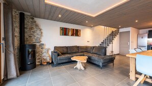 Una sala moderna con pared de piedra, un estufa de leña negra, un sofá seccional gris, una mesa de centro de madera y una escalera que sube a un nivel superior.
