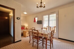 A dining area with a table set for four, wooden chairs, a chandelier, a television on the wall, and a door leading to another room.