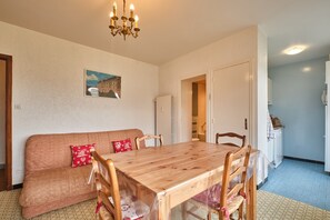 Un coin repas avec une table et des chaises en bois, un canapé avec des coussins rouges, un lustre et un espace cuisine équipé d’un réfrigérateur.