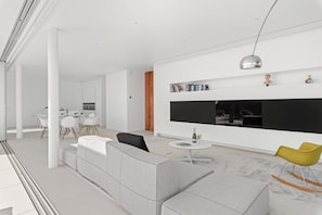 A modern living room with a sofa, a dining area with chairs, a kitchen, and a large black TV on the wall.