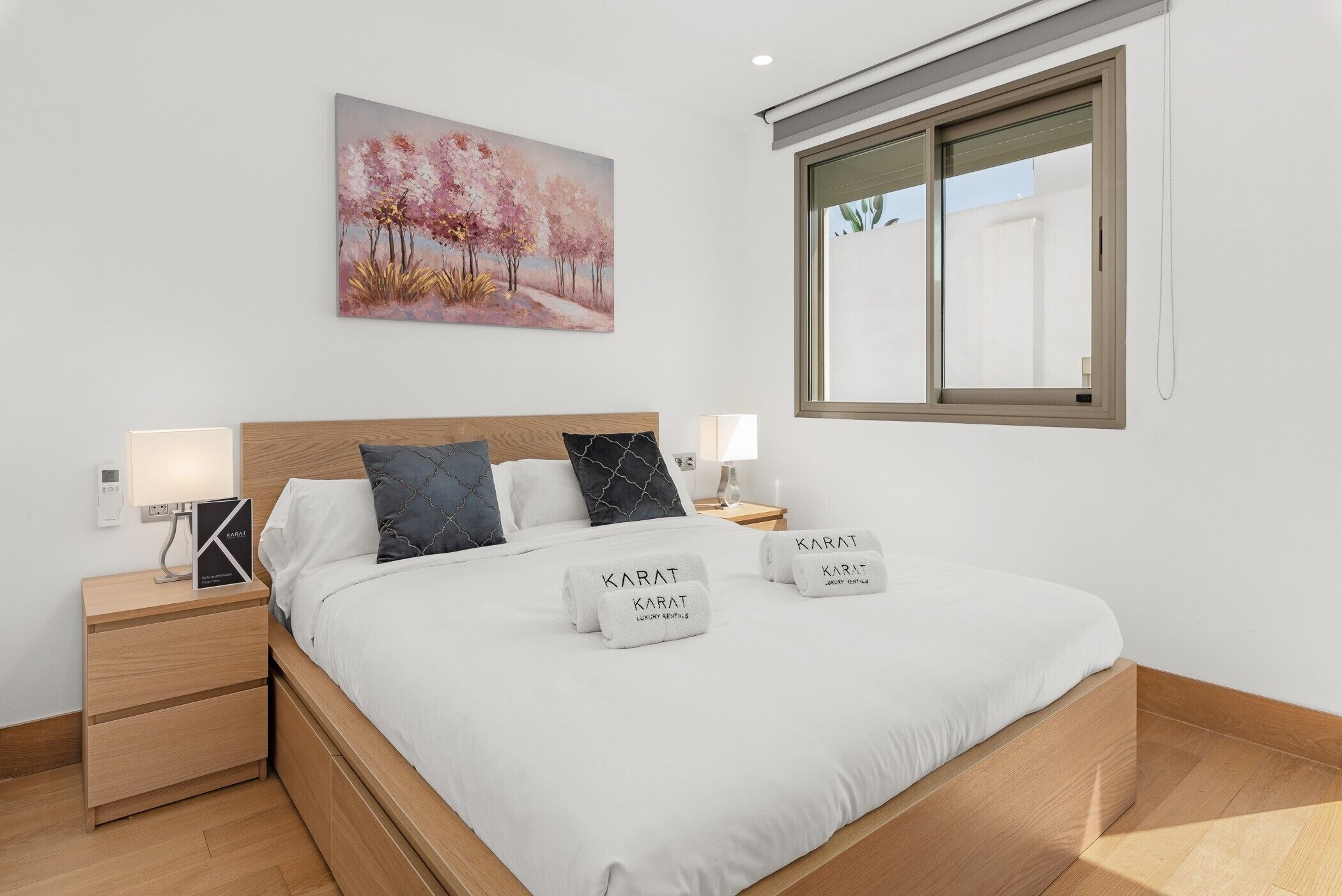 A neatly made bed with two towels, a bedside table with a lamp, a painting on the wall, and a window with blinds.