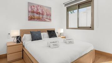 A neatly made bed with two towels, a bedside table with a lamp, a painting on the wall, and a window with blinds.