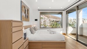 Une chambre à coucher avec un lit, des commodes en bois, un tableau, une fenêtre donnant sur des immeubles et un balcon.